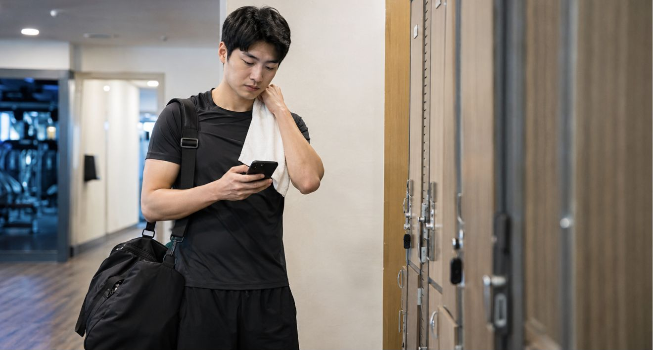 헬스장 라커룸 앞에서 운동 가방을 멘 사람이 휴대폰 화면을 잠시 내려다보는 장면