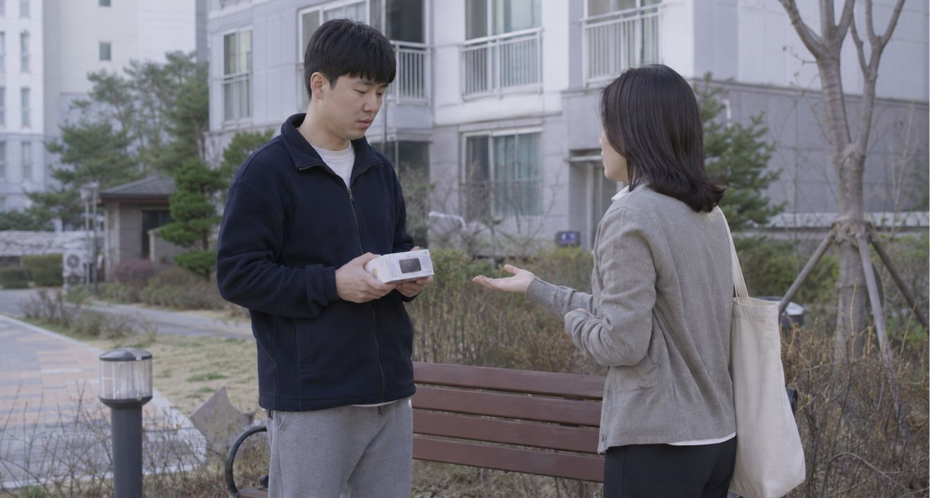 아파트 단지 앞 벤치에서 중고 물건을 들고 난감하게 서 있는 판매자와 구매자의 거래 장면