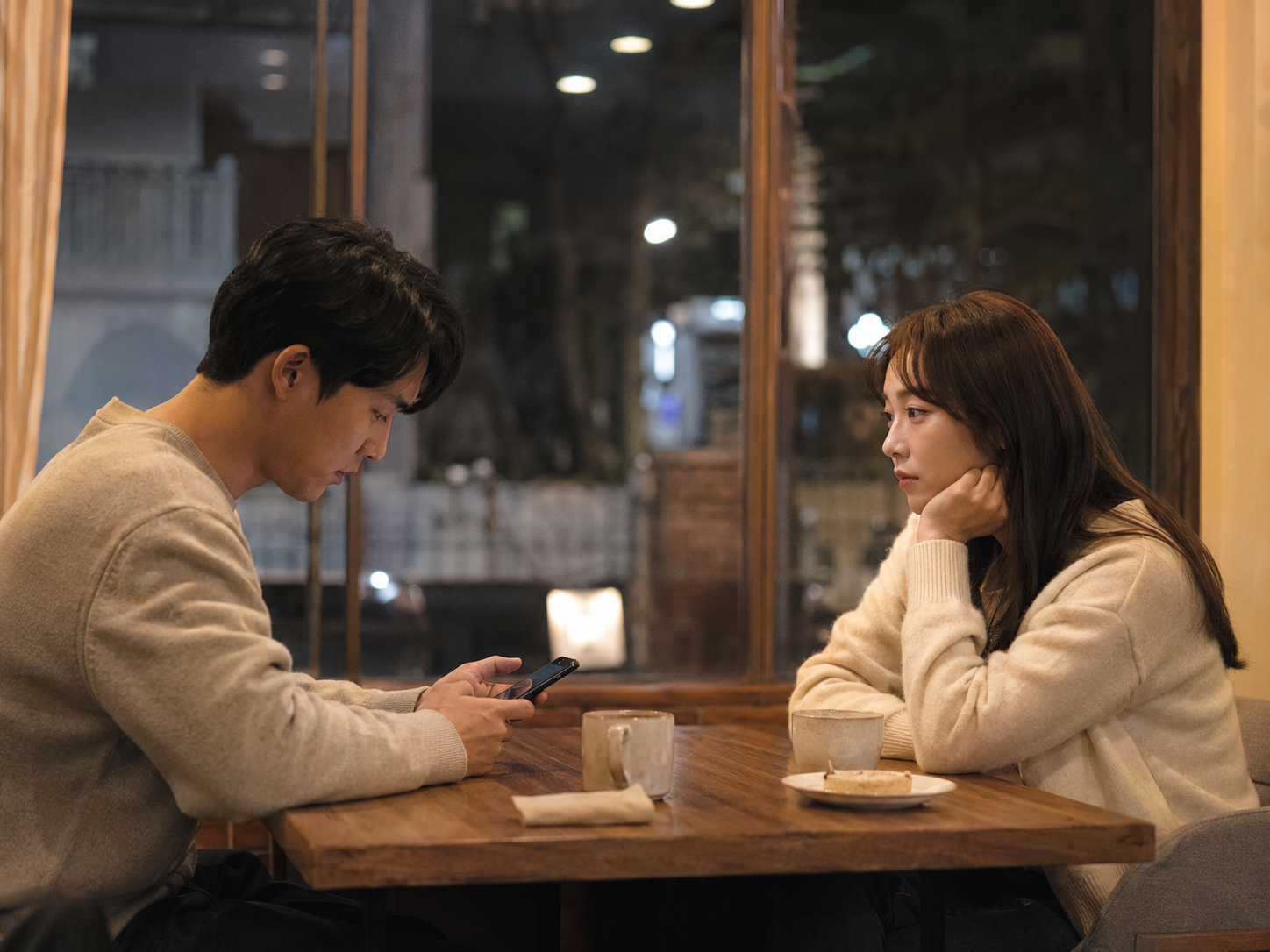 서울의 카페에서 한 사람은 휴대폰을 내려다보고 다른 한 사람은 조용히 표정을 굳힌 한국인 남녀